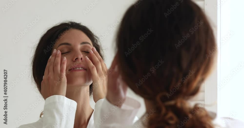 Head shot close up mirror reflection smiling young beautiful woman in bathrobe making facial massage satisfied with perfect healthy skin condition, involved in daily morning skincare routine. Head shot close up mirror reflection smiling young beautiful woman in bathrobe making facial massage satisfied with perfect healthy skin condition, involved in daily morning skincare routine.