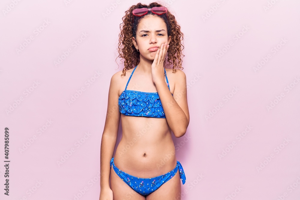 Naklejka premium Beautiful kid girl with curly hair wearing bikini and sunglasses touching mouth with hand with painful expression because of toothache or dental illness on teeth. dentist