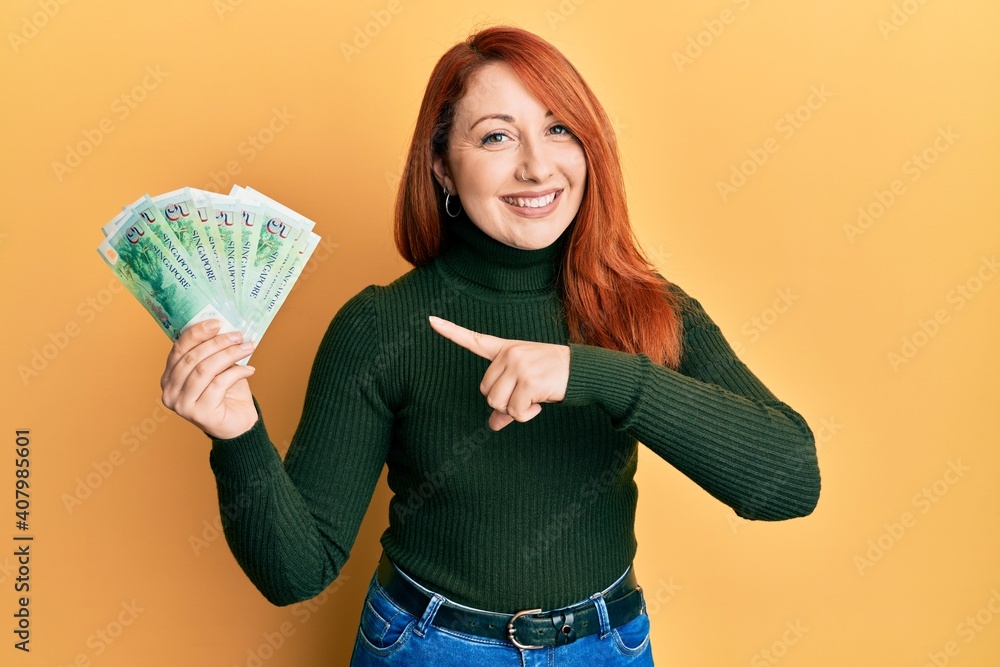 Beautiful redhead woman holding 5 singapore dollars banknotes smiling happy pointing with hand and finger Beautiful redhead woman holding 5 singapore dollars banknotes smiling happy pointing with hand and finger