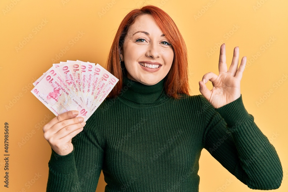 Beautiful redhead woman holding 10 turkish lira banknotes doing ok sign with fingers, smiling friendly gesturing excellent symbol Beautiful redhead woman holding 10 turkish lira banknotes doing ok sign with fingers, smiling friendly gesturing excellent symbol