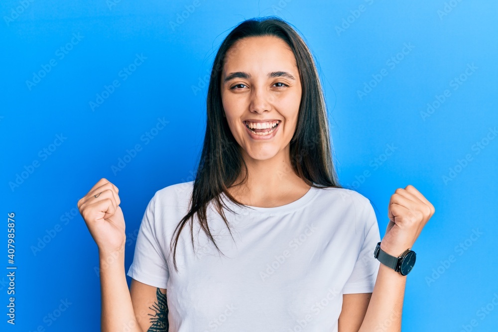 Young hispanic woman wearing casual white t shirt celebrating surprised and amazed for success with arms raised and open eyes. winner concept. Young hispanic woman wearing casual white t shirt celebrating surprised and amazed for success with arms raised and open eyes. winner concept.