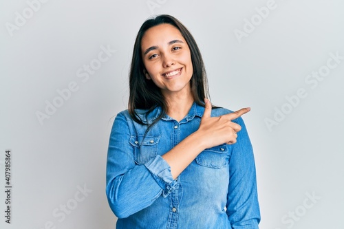 Young hispanic woman wearing casual denim jacket cheerful with a smile of face pointing with hand and finger up to the side with happy and natural expression on face