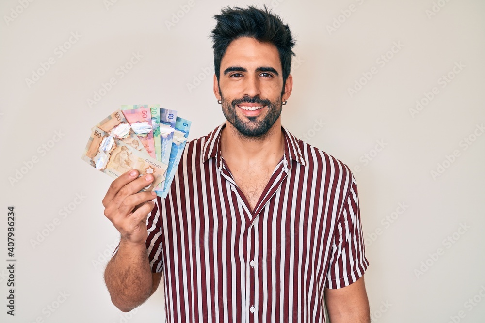 Handsome hispanic man with beard holding canadian dollars looking positive and happy standing and smiling with a confident smile showing teeth Handsome hispanic man with beard holding canadian dollars looking positive and happy standing and smiling with a confident smile showing teeth