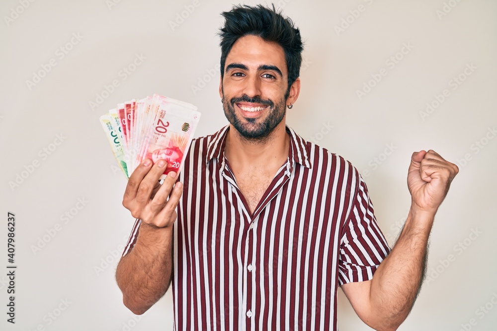 Handsome hispanic man with beard holding israel shekels screaming proud, celebrating victory and success very excited with raised arm Handsome hispanic man with beard holding israel shekels screaming proud, celebrating victory and success very excited with raised arm