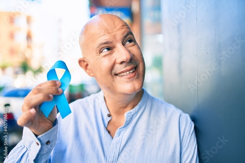 Middle age bald man smiling happy holding blue prostate cancer ribbon leaning on the wall at the city