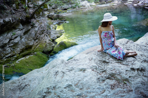 Fototapeta Naklejka Na Ścianę i Meble -  Young caucasian woman in a dress and hat resting on stones near a mountain river.