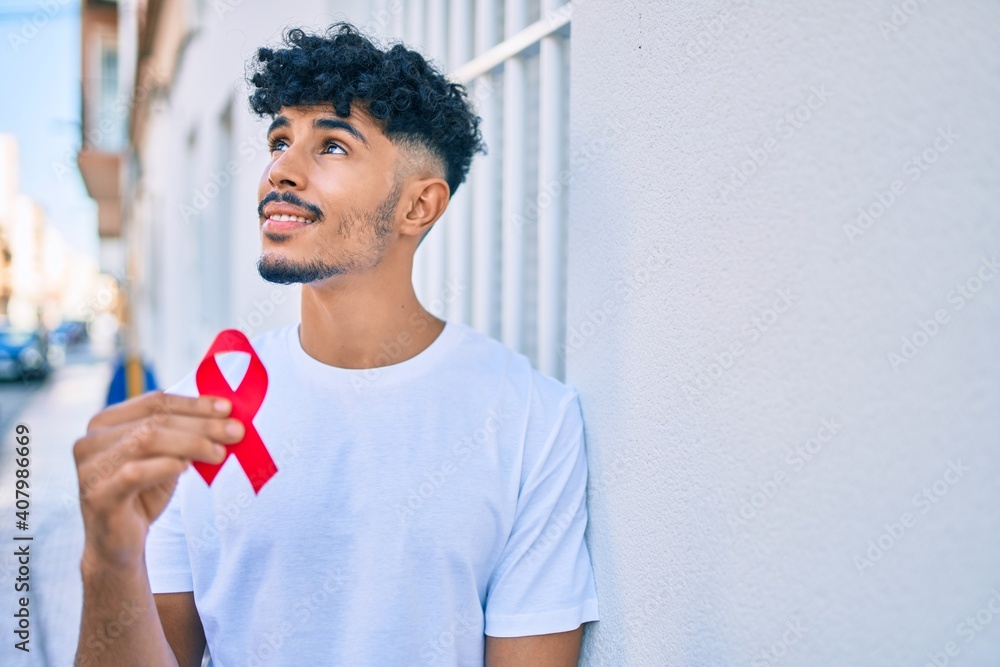 Young arab man smiling happy holding hiv awaraness red ribbon leaning on the wall. - obrazy, fototapety, plakaty 