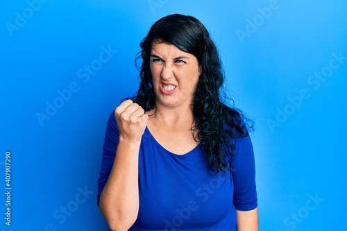Plus size brunette woman wearing casual blue shirt angry and mad raising fist frustrated and furious while shouting with anger. rage and aggressive concept.