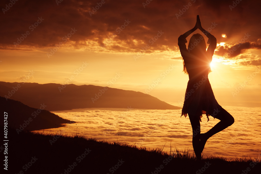 Young woman in dress doing yoga high in the mountains at dawn above the clouds.