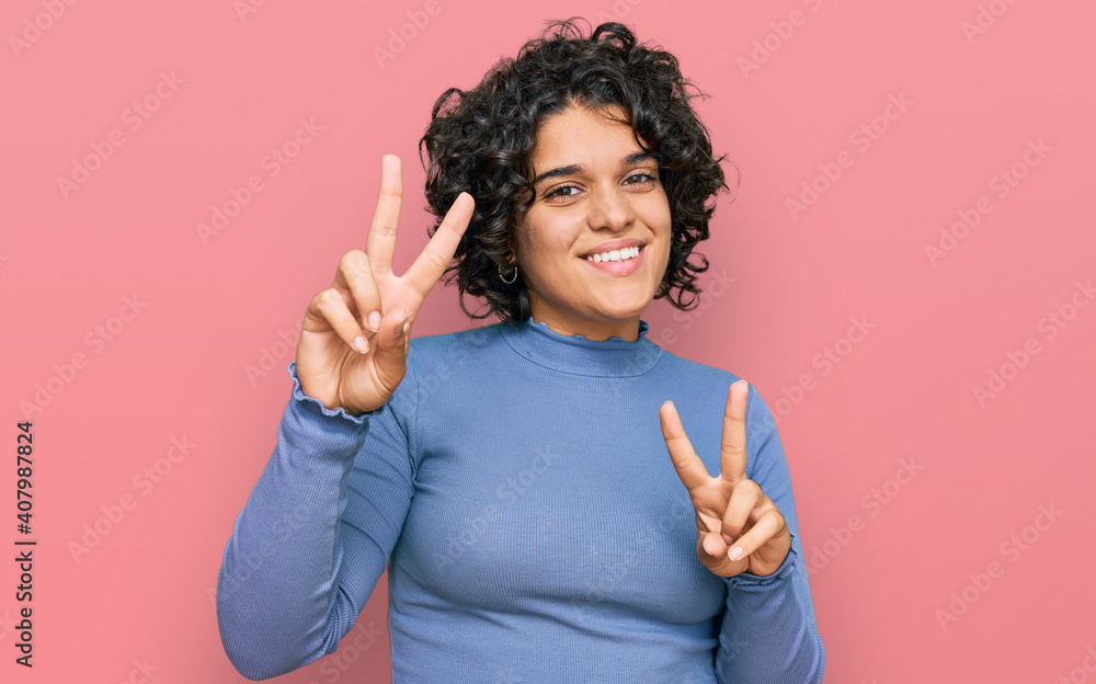 Young hispanic woman with curly hair wearing casual clothes smiling looking to the camera showing fingers doing victory sign. number two. Young hispanic woman with curly hair wearing casual clothes smiling looking to the camera showing fingers doing victory sign. number two.