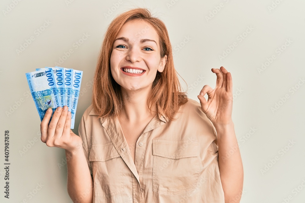 Obraz premium Young caucasian woman holding hungarian forint banknotes doing ok sign with fingers, smiling friendly gesturing excellent symbol