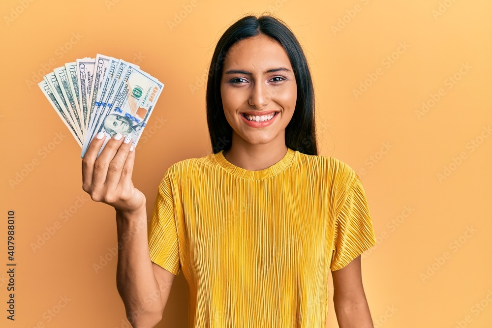 Young brunette woman holding dollars looking positive and happy standing and smiling with a confident smile showing teeth Young brunette woman holding dollars looking positive and happy standing and smiling with a confident smile showing teeth