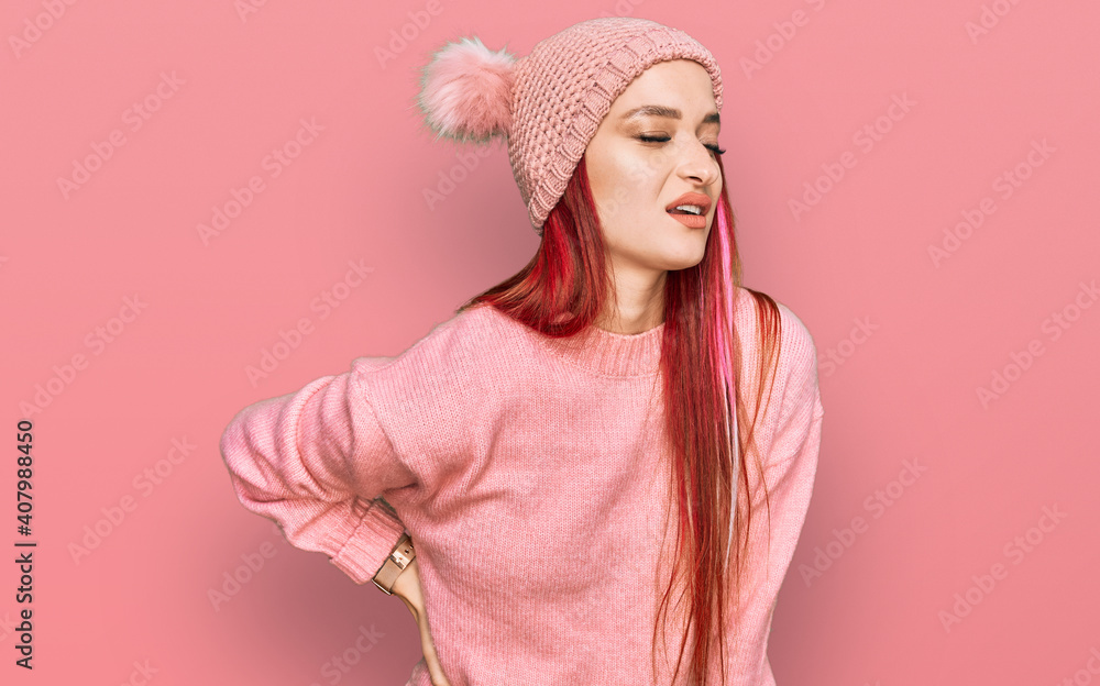 Young caucasian woman wearing casual clothes and wool cap suffering of backache, touching back with hand, muscular pain Young caucasian woman wearing casual clothes and wool cap suffering of backache, touching back with hand, muscular pain