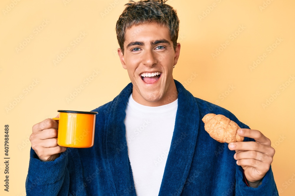Young handsome man wearing bathrobe eating breakfast holding coffee and croissant smiling and laughing hard out loud because funny crazy joke. Young handsome man wearing bathrobe eating breakfast holding coffee and croissant smiling and laughing hard out loud because funny crazy joke.