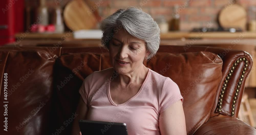 Older grey-haired female sit alone on leather couch at home with tablet, enjoy easy usage of modern wireless device, buy on e-commerce websites, search information, spend weekend on internet concept Older grey-haired female sit alone on leather couch at home with tablet, enjoy easy usage of modern wireless device, buy on e-commerce websites, search information, spend weekend on internet concept