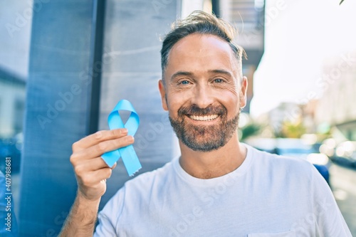Middle age handsome man smiling happy holding blue prostate cancer ribbon at the city.