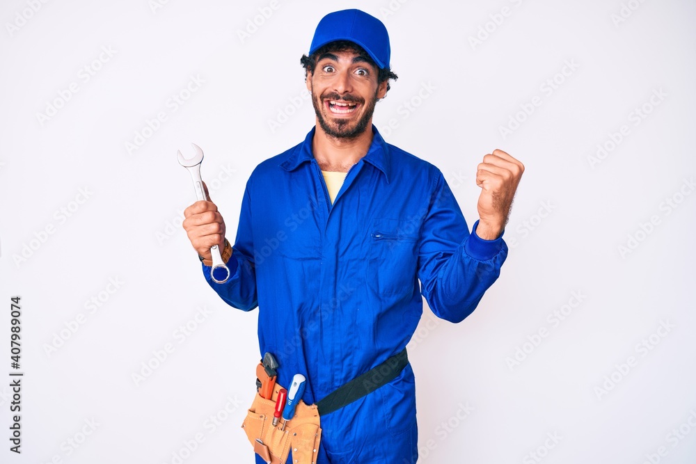 Handsome young man with curly hair and bear wearing builder jumpsuit uniform and holding wrench screaming proud, celebrating victory and success very excited with raised arms