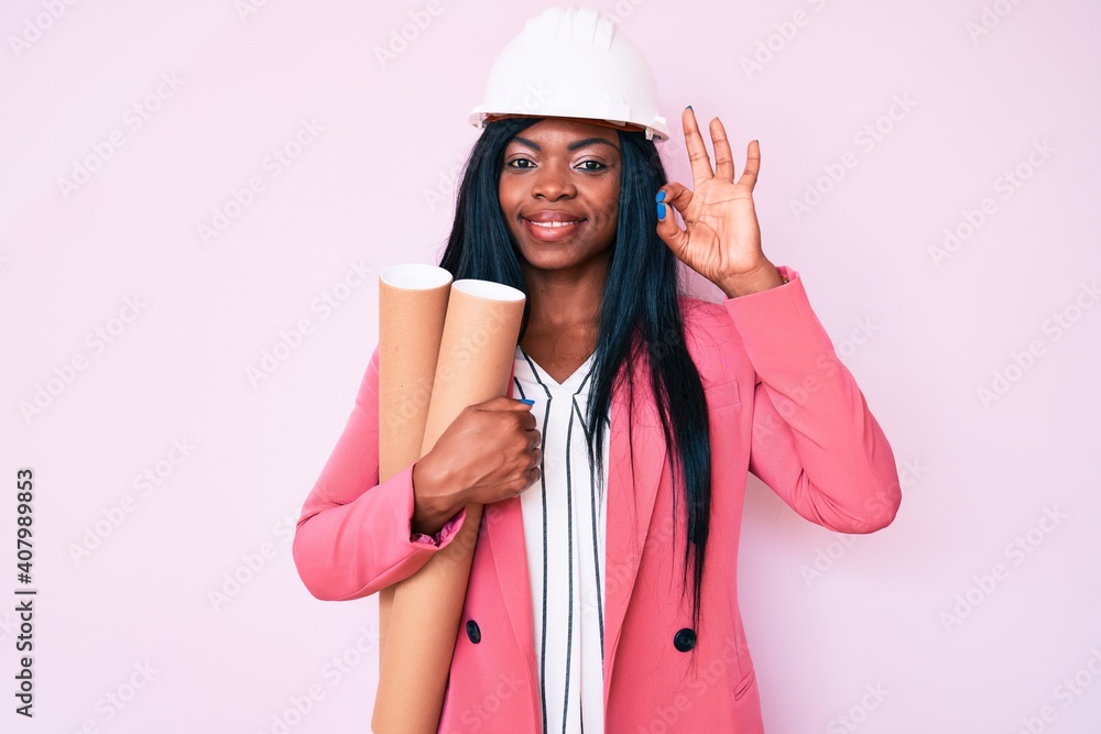 Young african american woman wearing architect hardhat holding blueprints doing ok sign with fingers, smiling friendly gesturing excellent symbol Young african american woman wearing architect hardhat holding blueprints doing ok sign with fingers, smiling friendly gesturing excellent symbol