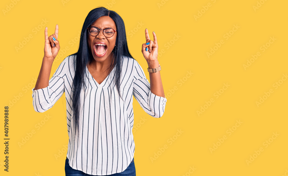 Young african american woman wearing casual clothes and glasses shouting with crazy expression doing rock symbol with hands up. music star. heavy music concept. Young african american woman wearing casual clothes and glasses shouting with crazy expression doing rock symbol with hands up. music star. heavy music concept.