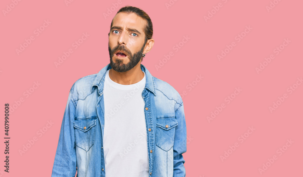 Attractive man with long hair and beard wearing casual denim jacket in shock face, looking skeptical and sarcastic, surprised with open mouth Attractive man with long hair and beard wearing casual denim jacket in shock face, looking skeptical and sarcastic, surprised with open mouth