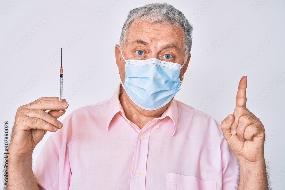 Senior grey-haired man wearing medical mask holding syringe surprised with an idea or question pointing finger with happy face, number one Senior grey-haired man wearing medical mask holding syringe surprised with an idea or question pointing finger with happy face, number one