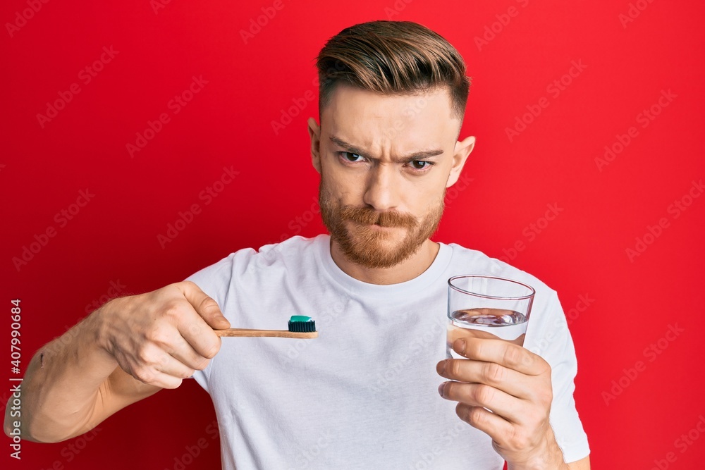 Young redhead man holding toothbrush with toothpaste and glass of water skeptic and nervous, frowning upset because of problem. negative person. Young redhead man holding toothbrush with toothpaste and glass of water skeptic and nervous, frowning upset because of problem. negative person.