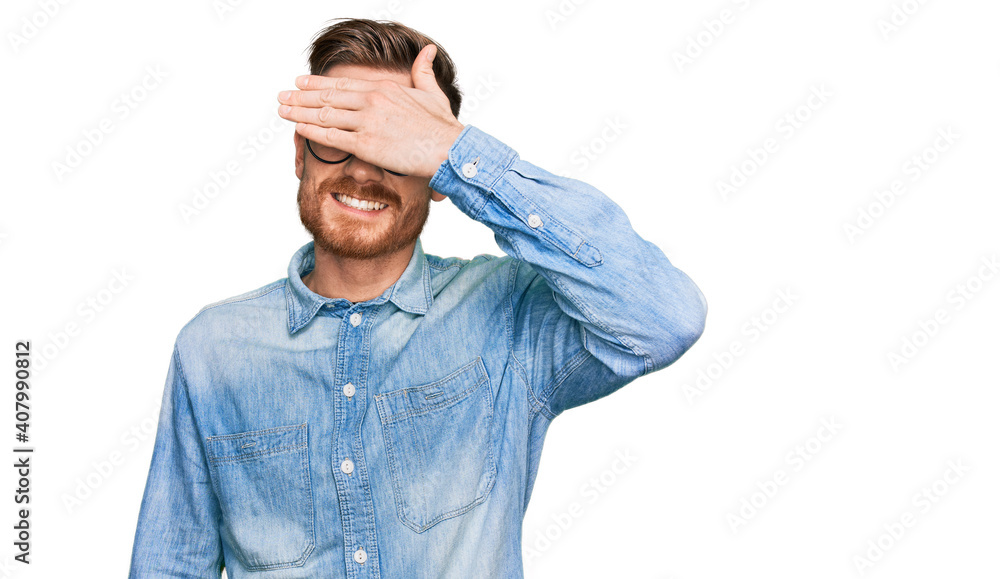Young redhead man wearing casual denim shirt smiling and laughing with hand on face covering eyes for surprise. blind concept. Young redhead man wearing casual denim shirt smiling and laughing with hand on face covering eyes for surprise. blind concept.