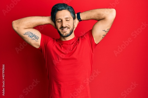 Young hispanic man wearing casual red t shirt relaxing and stretching, arms and hands behind head and neck smiling happy