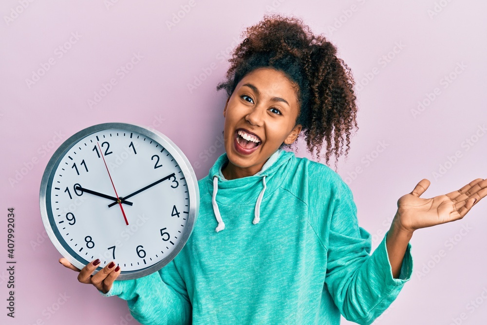 Beautiful african american woman with afro hair holding big clock celebrating achievement with happy smile and winner expression with raised hand Beautiful african american woman with afro hair holding big clock celebrating achievement with happy smile and winner expression with raised hand