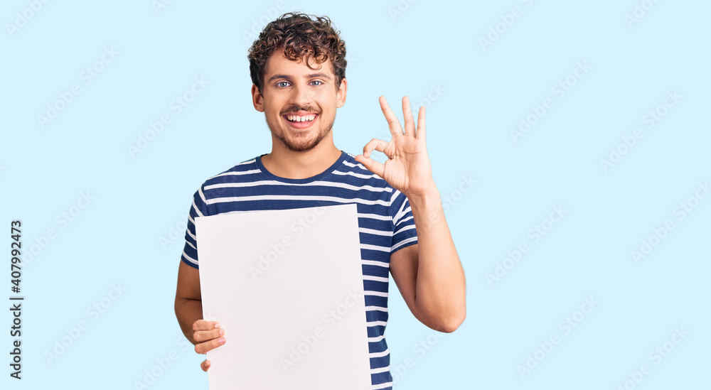 Young handsome man with curly hair holding blank empty banner doing ok sign with fingers, smiling friendly gesturing excellent symbol Young handsome man with curly hair holding blank empty banner doing ok sign with fingers, smiling friendly gesturing excellent symbol