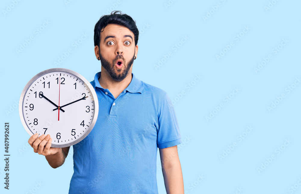 Young hispanic man holding big clock scared and amazed with open mouth for surprise, disbelief face