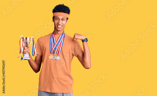 Young african american man holding champion trophy wearing medals pointing finger to one self smiling happy and proud