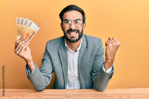 Obraz na płótnie Young hispanic man holding norwegian krone banknotes sitting on the table scream