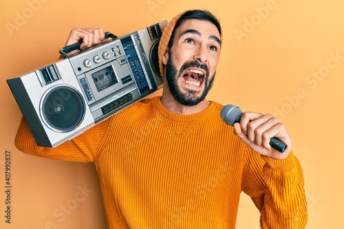 Young hispanic man holding boombox, listening to music singing with microphone angry and mad screaming frustrated and furious, shouting with anger looking up.