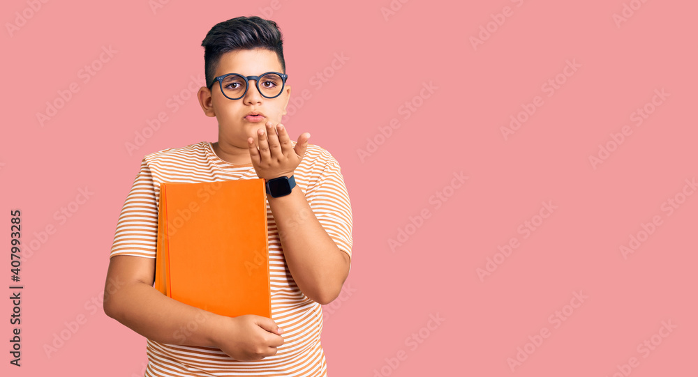 Little boy kid holding book wearing glasses looking at the camera blowing a kiss with hand on air being lovely and sexy. love expression. Little boy kid holding book wearing glasses looking at the camera blowing a kiss with hand on air being lovely and sexy. love expression.