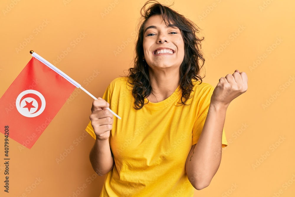 Young hispanic woman holding tunisia flag screaming proud, celebrating victory and success very excited with raised arm Young hispanic woman holding tunisia flag screaming proud, celebrating victory and success very excited with raised arm