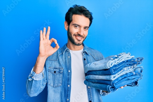 Young hispanic man holding stack of folded jeans doing ok sign with fingers, smiling friendly gesturing excellent symbol