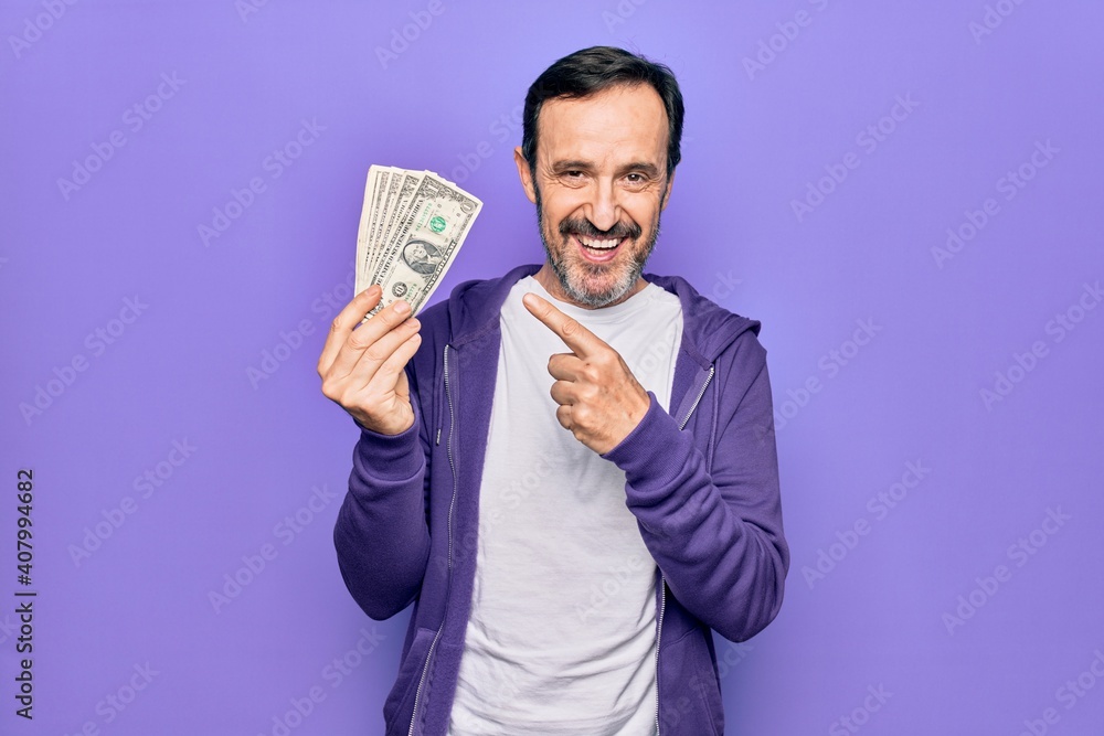 Middle age handsome man holding bunch of dollars banknotes over isolated purple background smiling happy pointing with hand and finger Middle age handsome man holding bunch of dollars banknotes over isolated purple background smiling happy pointing with hand and finger