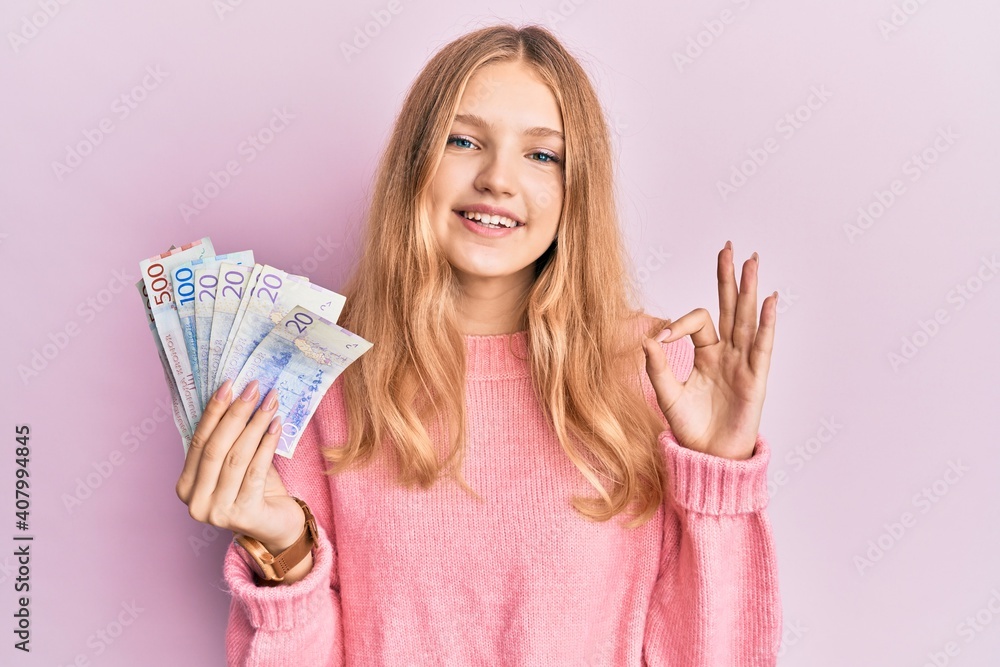 Beautiful young caucasian girl holding swedish krona banknotes doing ok sign with fingers, smiling friendly gesturing excellent symbol Beautiful young caucasian girl holding swedish krona banknotes doing ok sign with fingers, smiling friendly gesturing excellent symbol