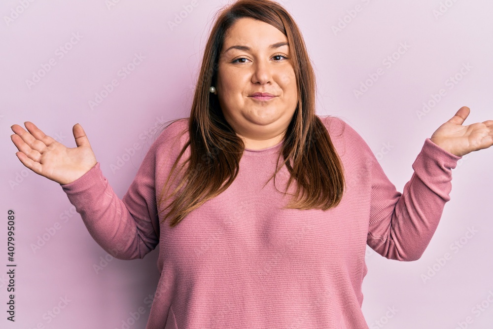Fototapeta premium Beautiful brunette plus size woman wearing casual winter sweater clueless and confused with open arms, no idea and doubtful face.