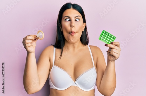 Beautiful brunette woman holding condom and birth control pills making fish face with mouth and squinting eyes, crazy and comical.