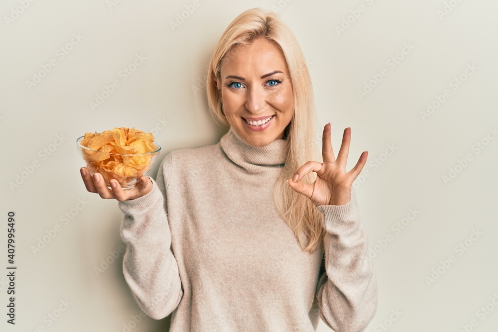 Young blonde woman holding bowl with potato chip doing ok sign with fingers, smiling friendly gesturing excellent symbol
