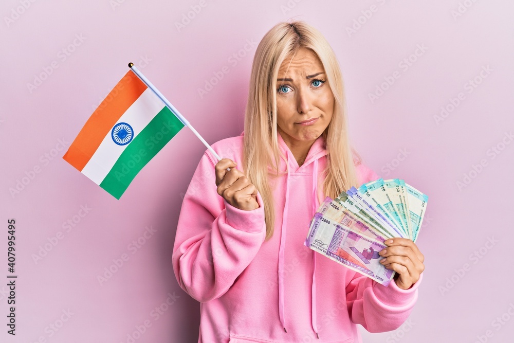 Young blonde woman holding india flag and rupee banknotes skeptic and nervous, frowning upset because of problem. negative person. Young blonde woman holding india flag and rupee banknotes skeptic and nervous, frowning upset because of problem. negative person.
