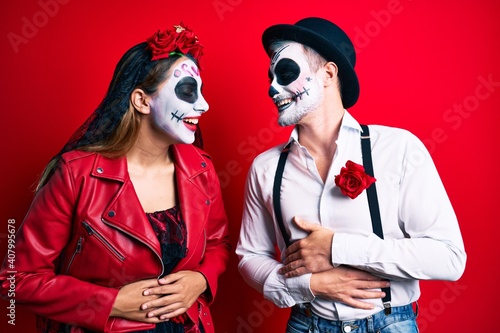 Couple wearing day of the dead costume over red smiling and laughing hard out loud because funny crazy joke with hands on body.