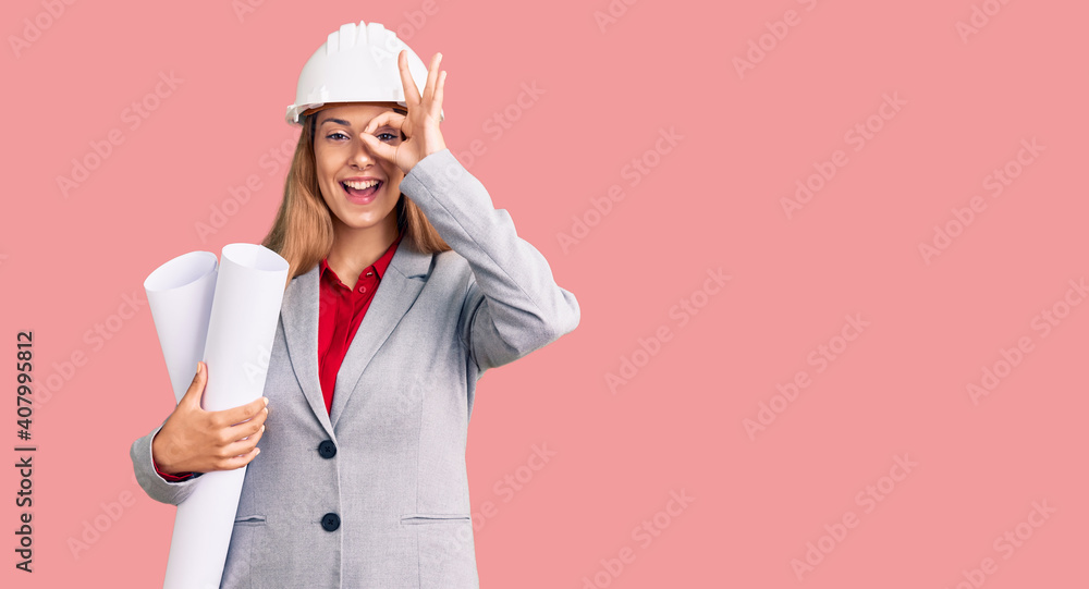 Beautiful young woman wearing architect hardhat holding build project smiling happy doing ok sign with hand on eye looking through fingers Beautiful young woman wearing architect hardhat holding build project smiling happy doing ok sign with hand on eye looking through fingers