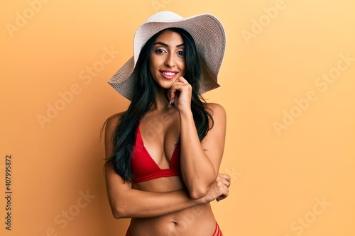 Beautiful hispanic woman wearing bikini and summer hat smiling looking confident at the camera with crossed arms and hand on chin. thinking positive.