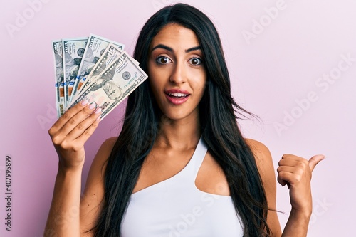 Beautiful hispanic woman holding dollars pointing thumb up to the side smiling happy with open mouth