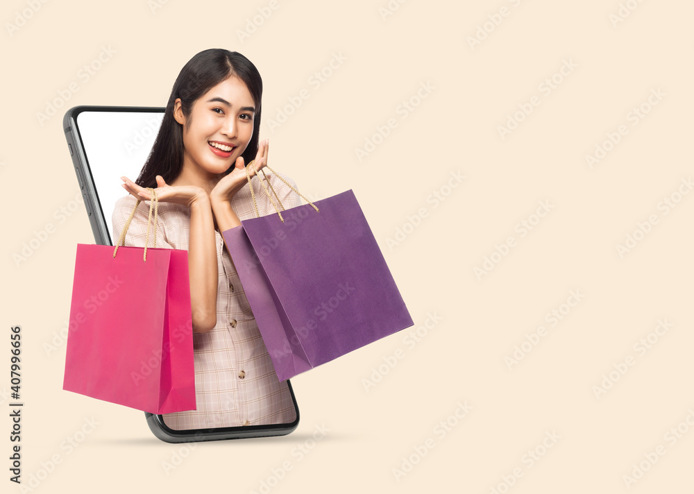 Young smiling beautiful asian woman making online shopping and holding shopping bags out through mobile phone isolated on beige background with clipping path.