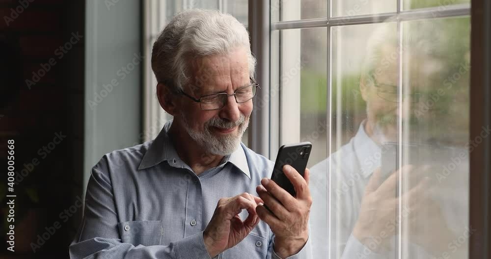 Happy aged bearded man in eyeglasses standing indoor lean on window using smartphone device. Easy on-line internet services for older generation, modern wireless gadget usage, mobile app, fun concept Happy aged bearded man in eyeglasses standing indoor lean on window using smartphone device. Easy on-line internet services for older generation, modern wireless gadget usage, mobile app, fun concept