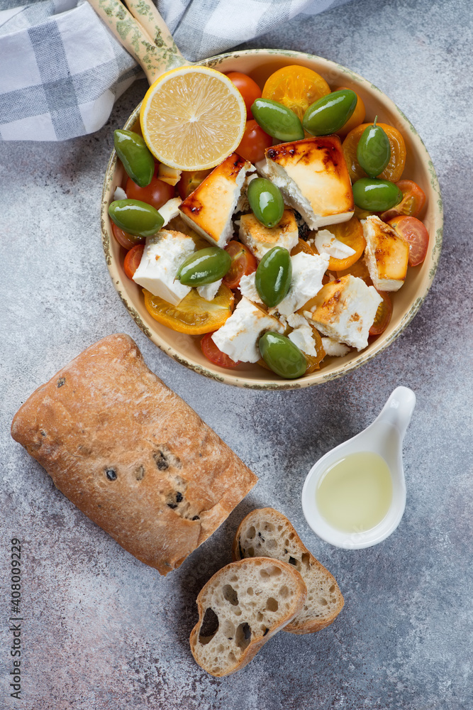Chunks of feta cheese baked with tomatoes and green olives and served with ciabatta, flatlay on a beige stone background